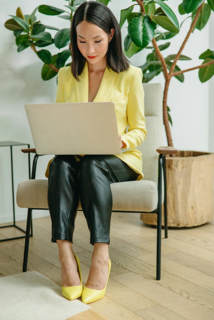 Asian woman in yellow blazer working on laptop indoors, showcasing modern digital nomad lifestyle.